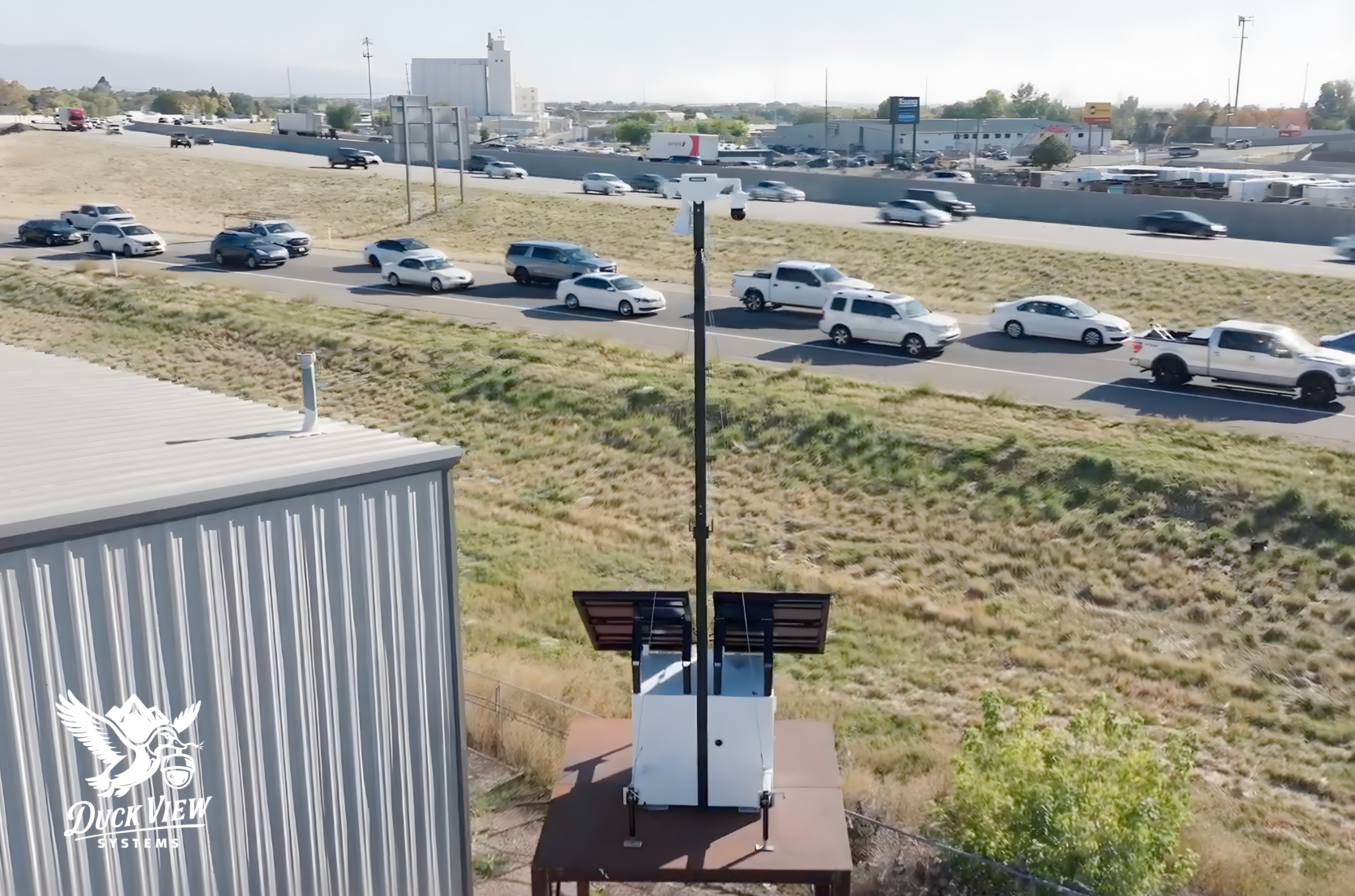 Mobile Camera Trailer Monitoring Traffic Near Industrial Site Mobile camera trailer with elevated mast and solar panels positioned near a highway and industrial buildings, providing wide-area surveillance, vehicle monitoring, and off-grid security coverage for commercial and roadside environments.